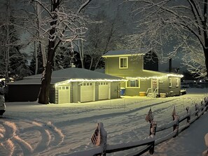 Een huis met twee verdiepingen, met garage, omgeven door met sneeuw bedekte bomen en een besneeuwde hekken.