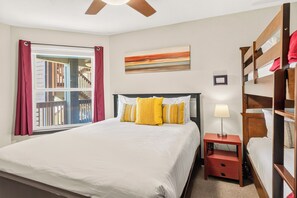 Ein Schlafzimmer mit einem Bett, einem Nachttisch, einer Lampe, einem Fenster mit Vorhängen, einem Gemälde und einem Etagenbett.