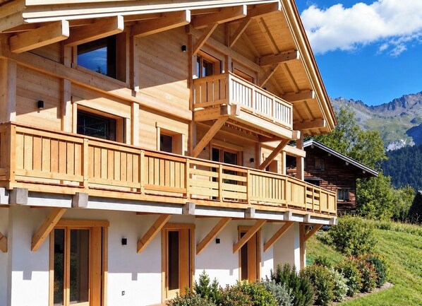Un chalet en bois à plusieurs étages, doté de balcons, qui se dresse sur un fond de paysage montagneux.