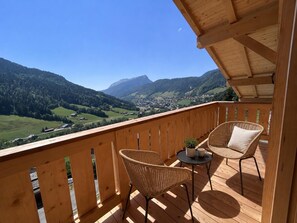 Une terrasse en bois, agrémentée de chaises en osier et d’une petite table, qui offre une vue sur une vallée avec des montagnes en arrière-plan.