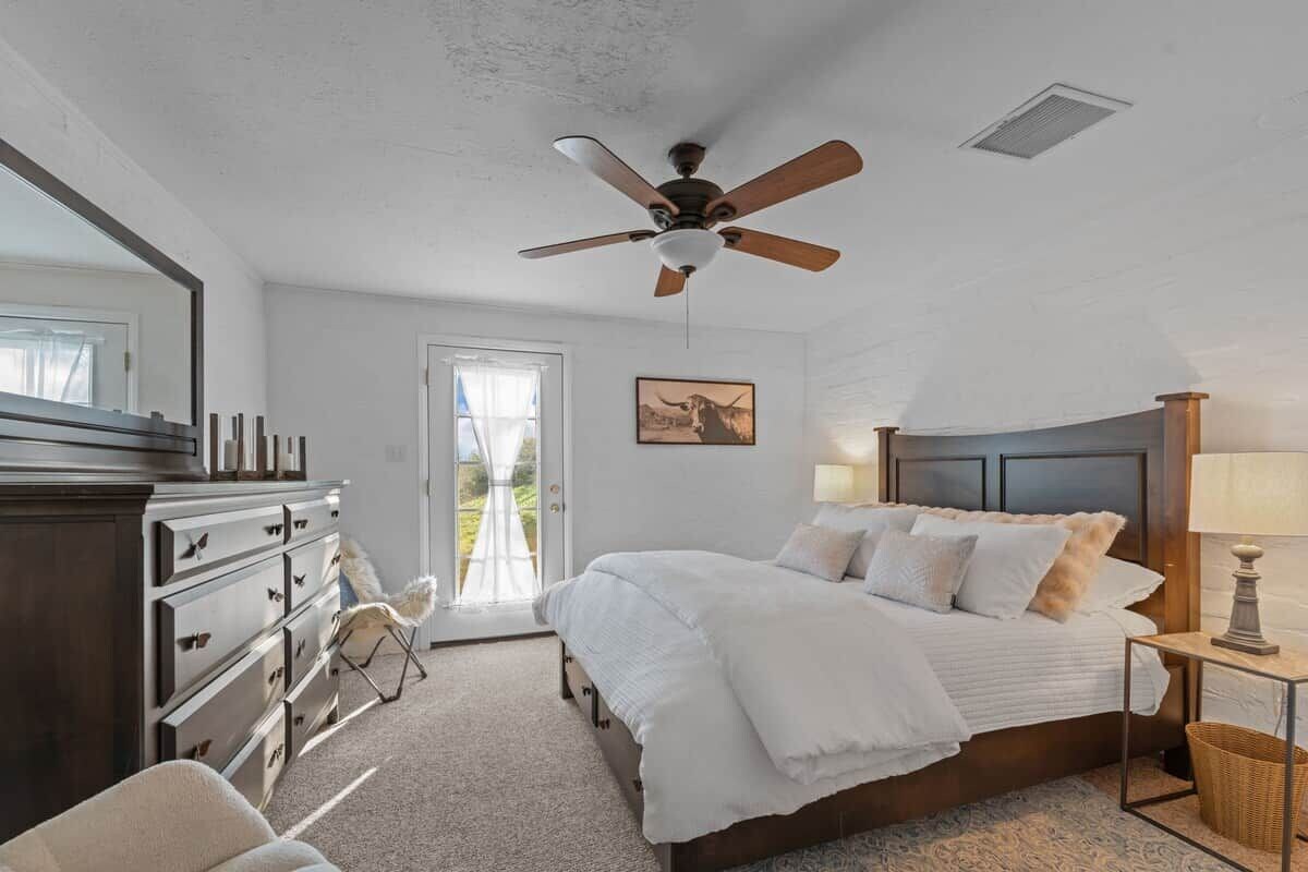 A bedroom with a bed, dresser, ceiling fan, and a window with a view.