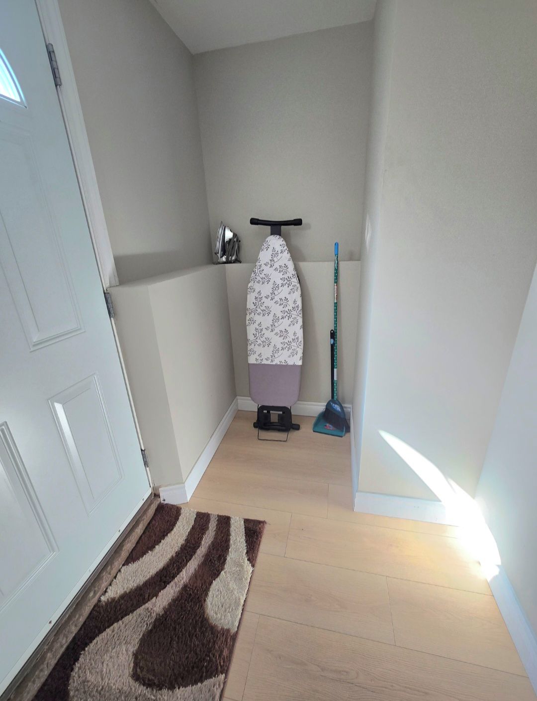 A hallway with a white door, a patterned ironing board, a broom, and a brown rug.