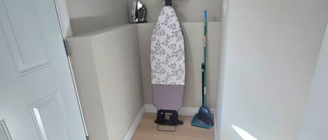 A hallway with a white door, a patterned ironing board, a broom, and a brown rug.