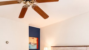 Ein Hotelzimmer mit einem Bett, einem Deckenventilator, einem Nachttisch mit Lampe und einer Wand mit einem Gemälde.