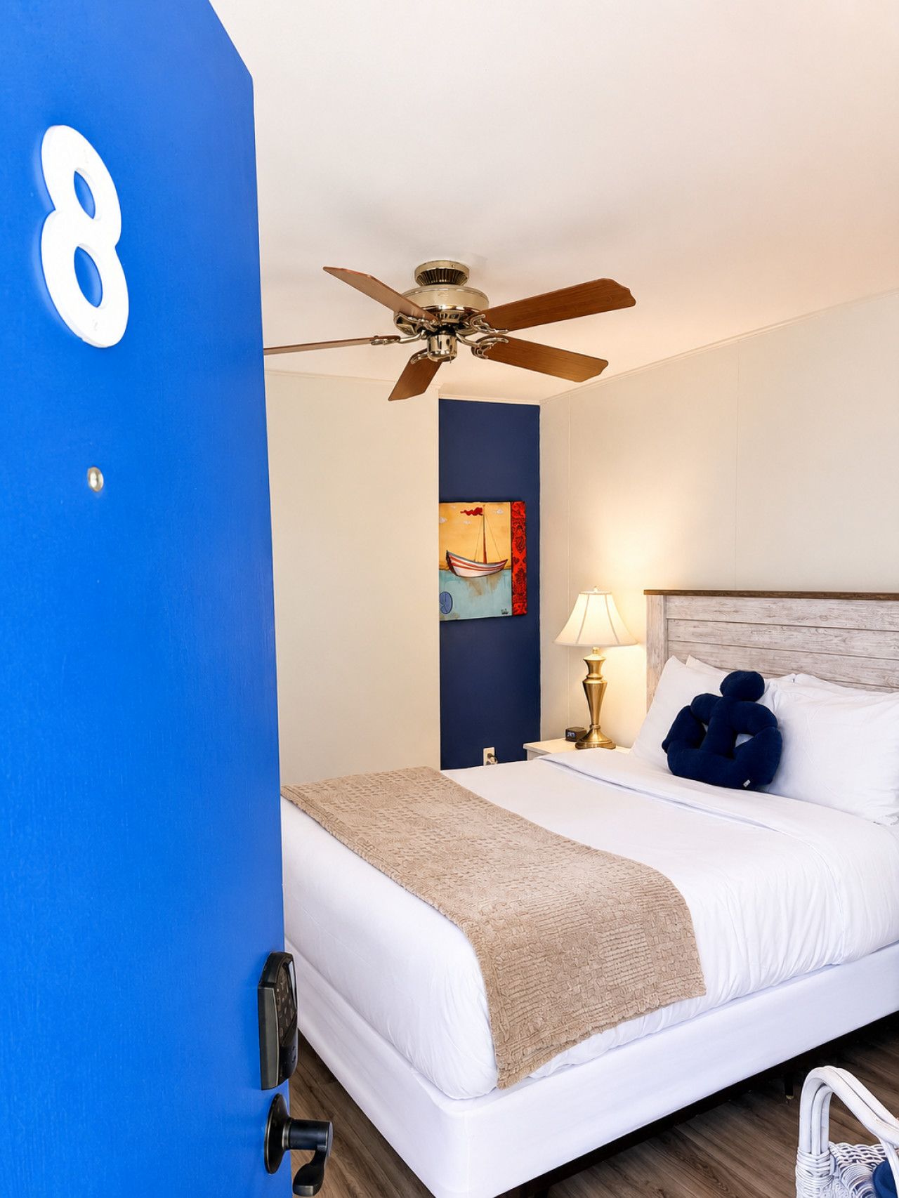 A hotel room with a bed, a ceiling fan, a painting, a nightstand with a lamp, and a blue wall with a number.