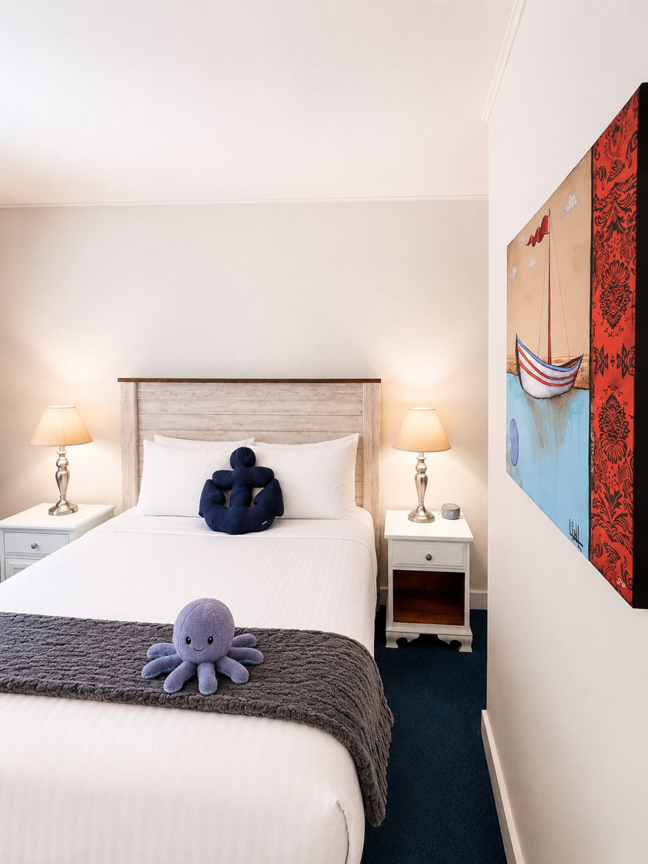 Una habitación de hotel con una cama, dos mesitas de noche, una lámpara, un peluche de pulpo y un cuadro en la pared.
