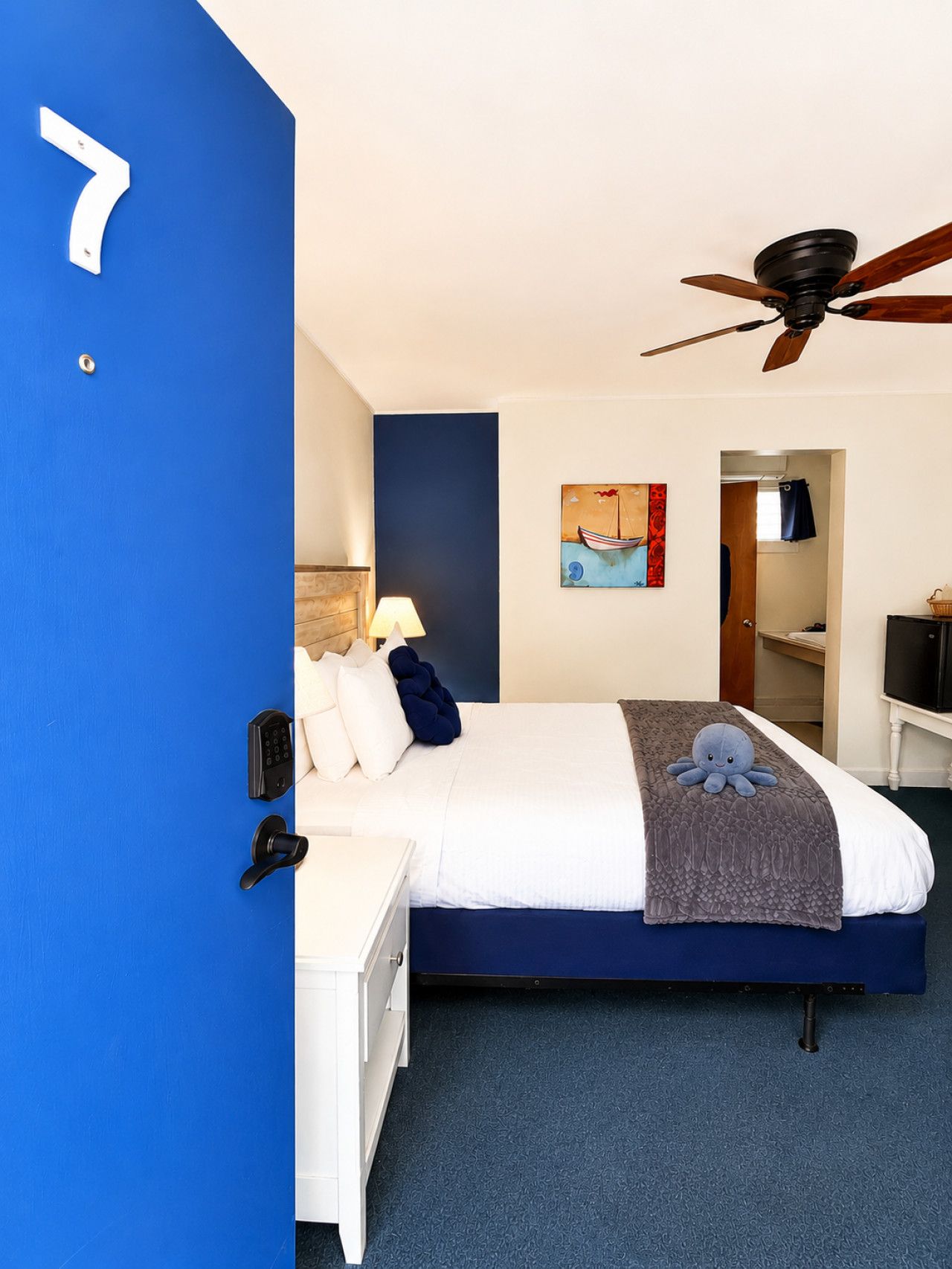 A hotel room with a bed, a ceiling fan, a painting, and a door with a number.