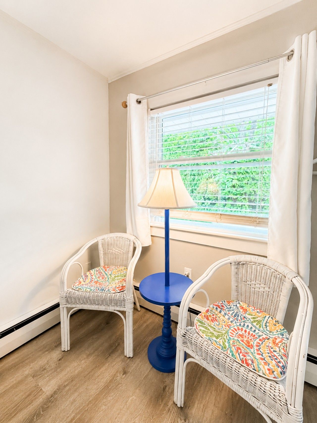 Ein Zimmer mit zwei Korbstühlen mit bunten Kissen, einem kleinen blauen Tisch und einer Lampe. Ein Fenster mit Jalousien und Blick auf Grün ist sichtbar.