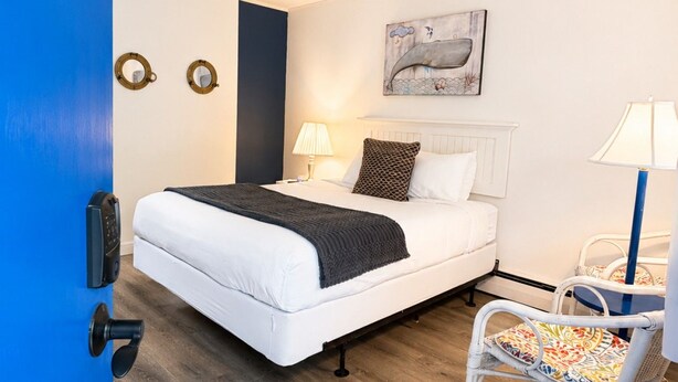 A bedroom with a bed, a ceiling fan, a wall-mounted artwork, two bedside lamps, and a chair with a colorful cushion. - Towne Motel Camben Room #6 (Camden)