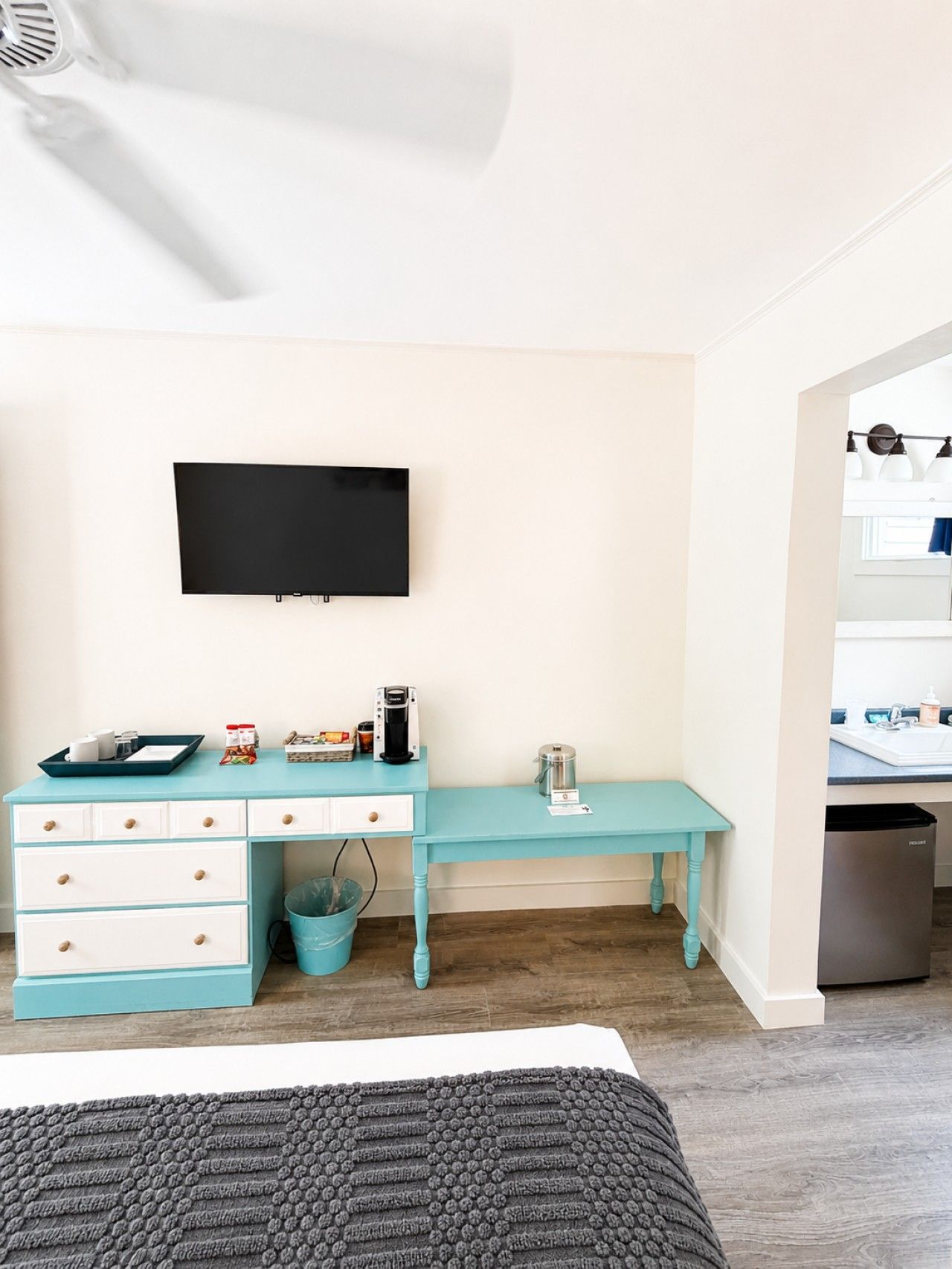 Un dormitorio con cama, escritorio con televisor, cafetera y cubo de basura.