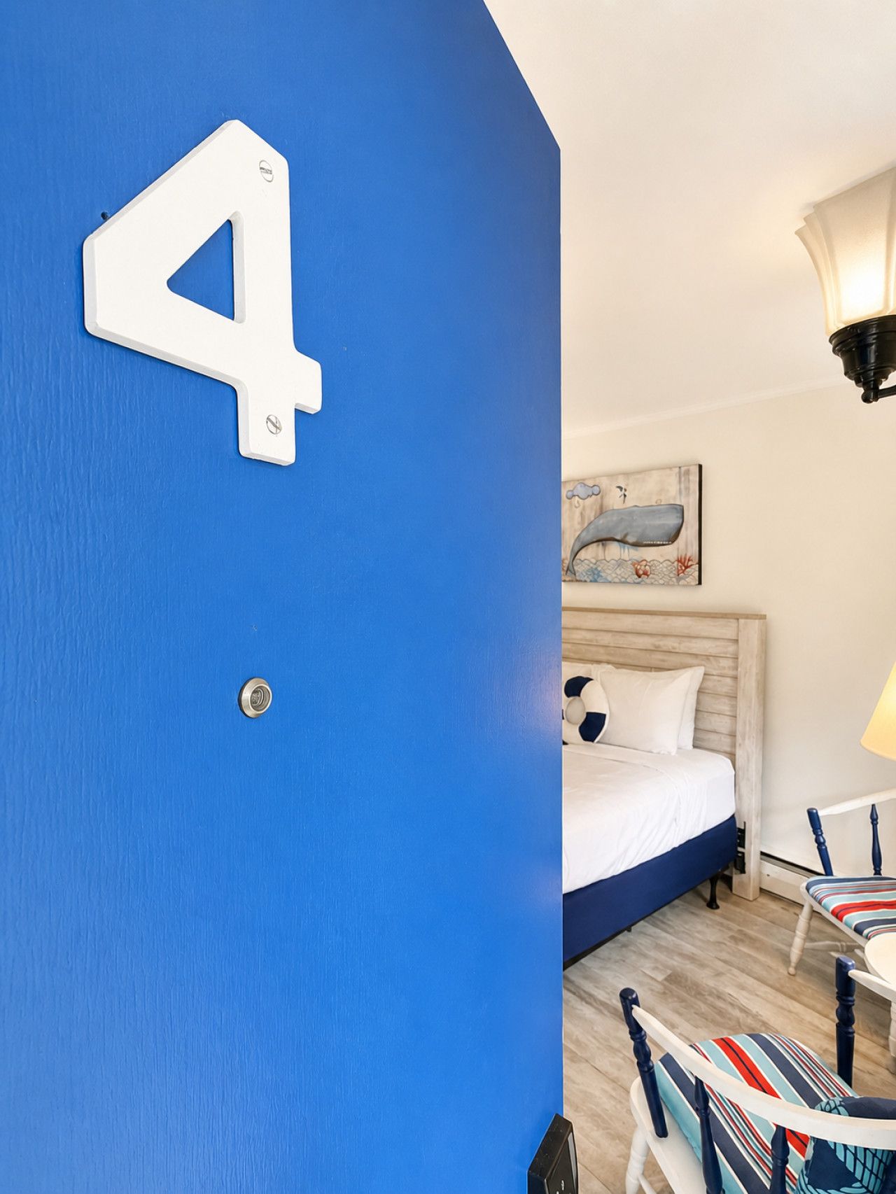 Ein Zimmer mit blauer Wand, einem Bett mit weißer Bettwäsche, einem Bild an der Wand, einer Lampe und einem Sessel mit gestreifter Sitzkissen.