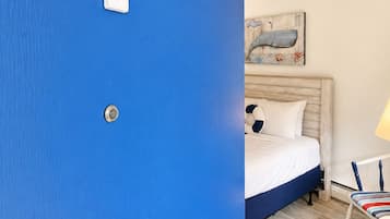 Una habitación con pared azul, cama con ropa de cama blanca, un cuadro en la pared, una lámpara y una silla con cojín a rayas.