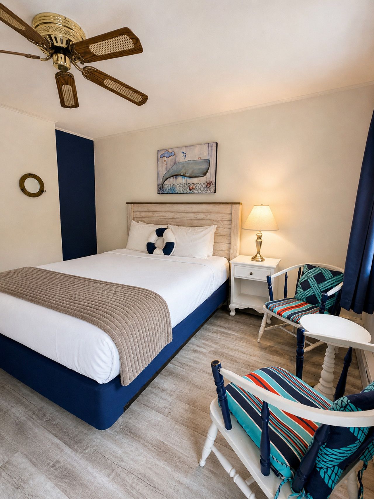Una habitación de hotel con cama, mesita de noche, lámpara, silla, mesa, ventilador de techo y un cuadro en la pared.