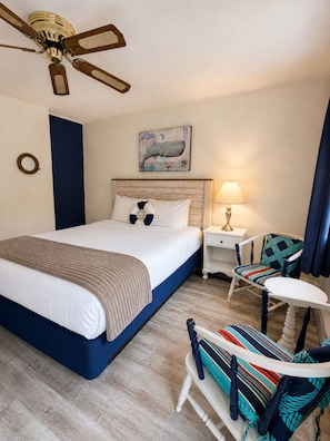 Habitació d'hotel amb un llit, una taula de nit, una làmpada, una cadira, una taula, un ventilador de sostre i un quadre penjat a la paret.