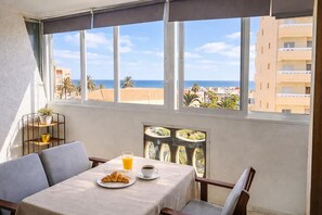 Un comedor con una mesa dispuesta para dos personas, vista a la playa por la ventana y una planta en maceta en un estante.