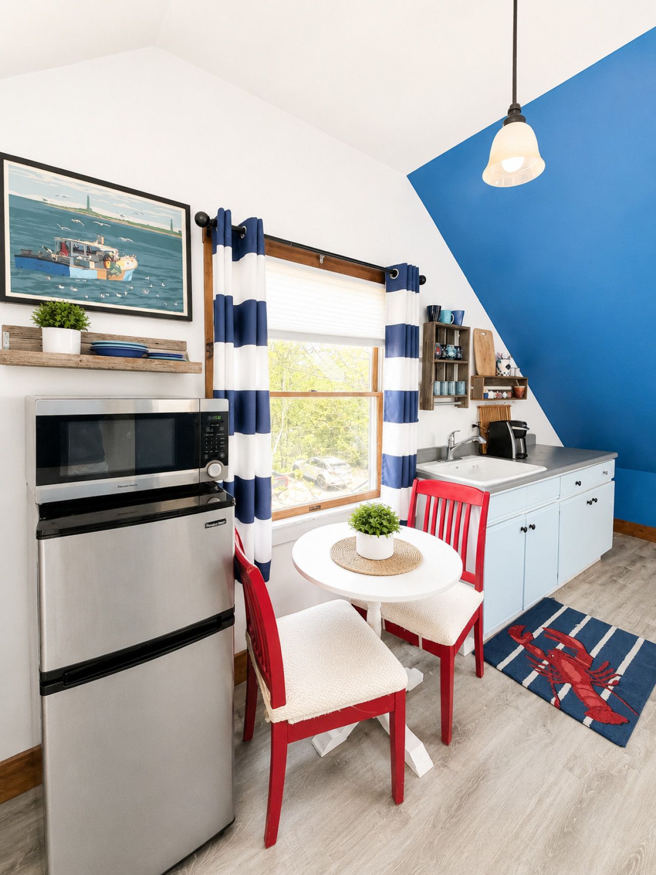 Cocina compacta con refrigerador, microondas y lavamanos. Hay un comedor con una mesa y sillas, y una ventana con cortinas. Un cuadro está en la pared, y hay una planta en la repisa.