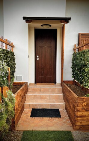 A wooden front door with a dark brown finish, flanked by two wooden planters with green foliage, leading to a set of stone steps. - Apartment in a villa with free parking and a private garden. (Abruzzo)