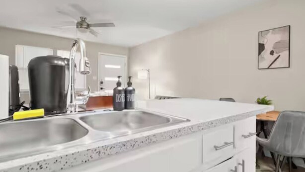 Una cocina moderna con encimera blanca, dos fregaderos, ventilador de techo y comedor con mesa y sillas.