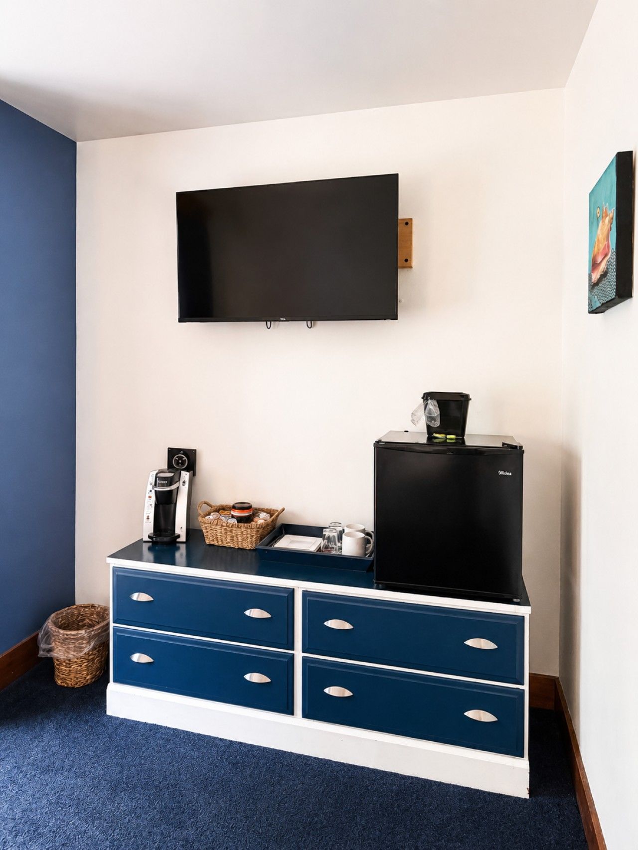 Habitación con una televisión de pantalla plana montada en la pared, cafetera, refrigerador y un mueble azul con cajones.
