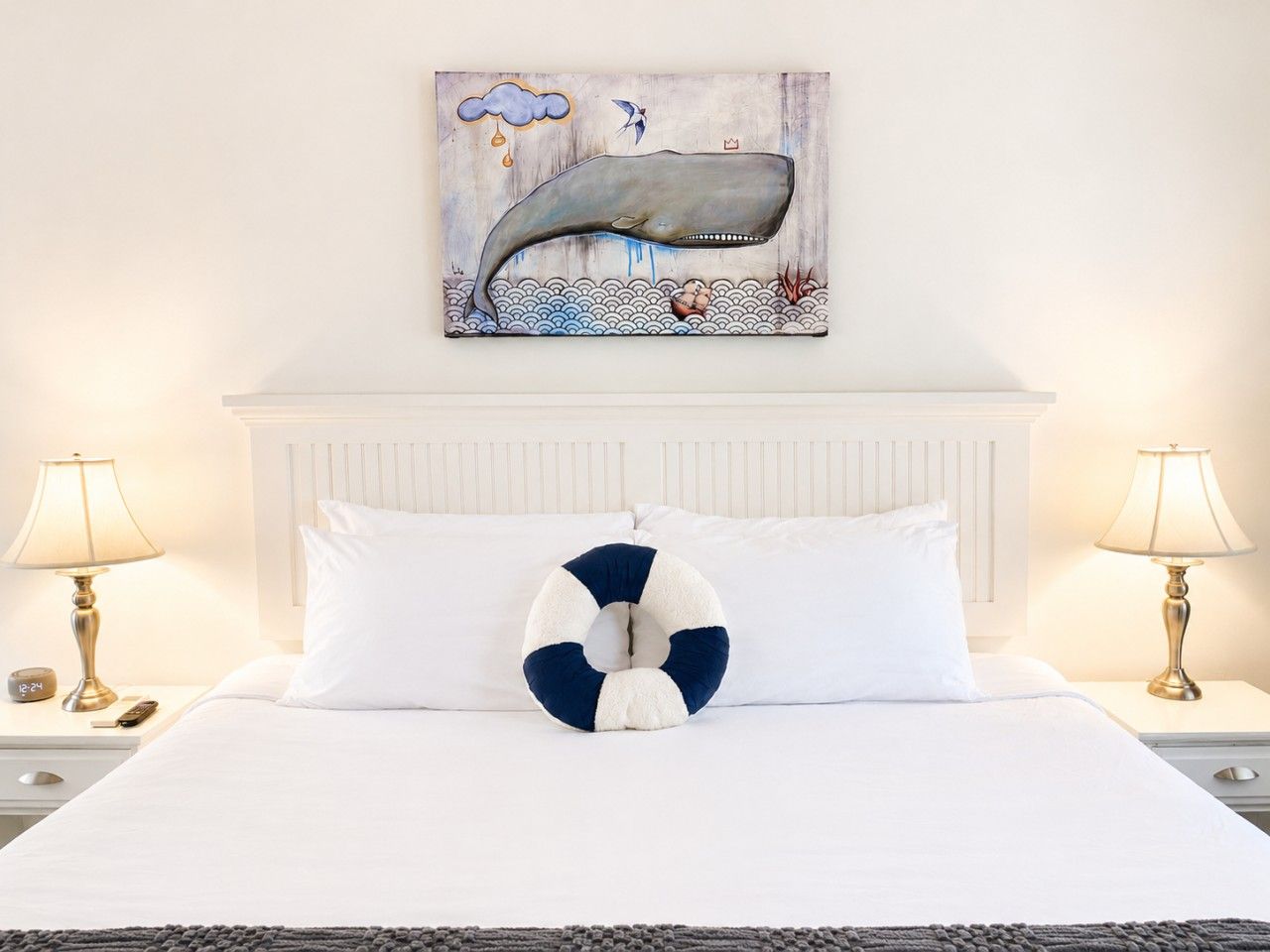 Una habitación de hotel con una cama, ropa de cama blanca, un cojín con forma de boya salvavidas, una obra de arte colgada en la pared de una ballena y dos lámparas de noche.