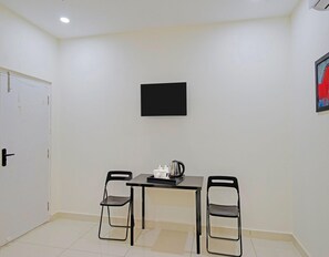 Um quarto pequeno com porta branca, mesa de canto com moldura preta e duas cadeiras, chaleira e quadro na parede.