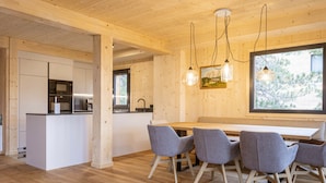 A modern kitchen with white cabinets and a wooden island, a dining area with a wooden table and grey chairs, and a large window with a view of trees. - Superior Chalet #23 With Sauna & Hot Tub (Stadl-Predlitz)