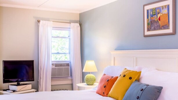 A hotel room with a bed, a television, a window with curtains, a lamp, and a painting on the wall. - Towne Motel Camben Room #16 (Camden)