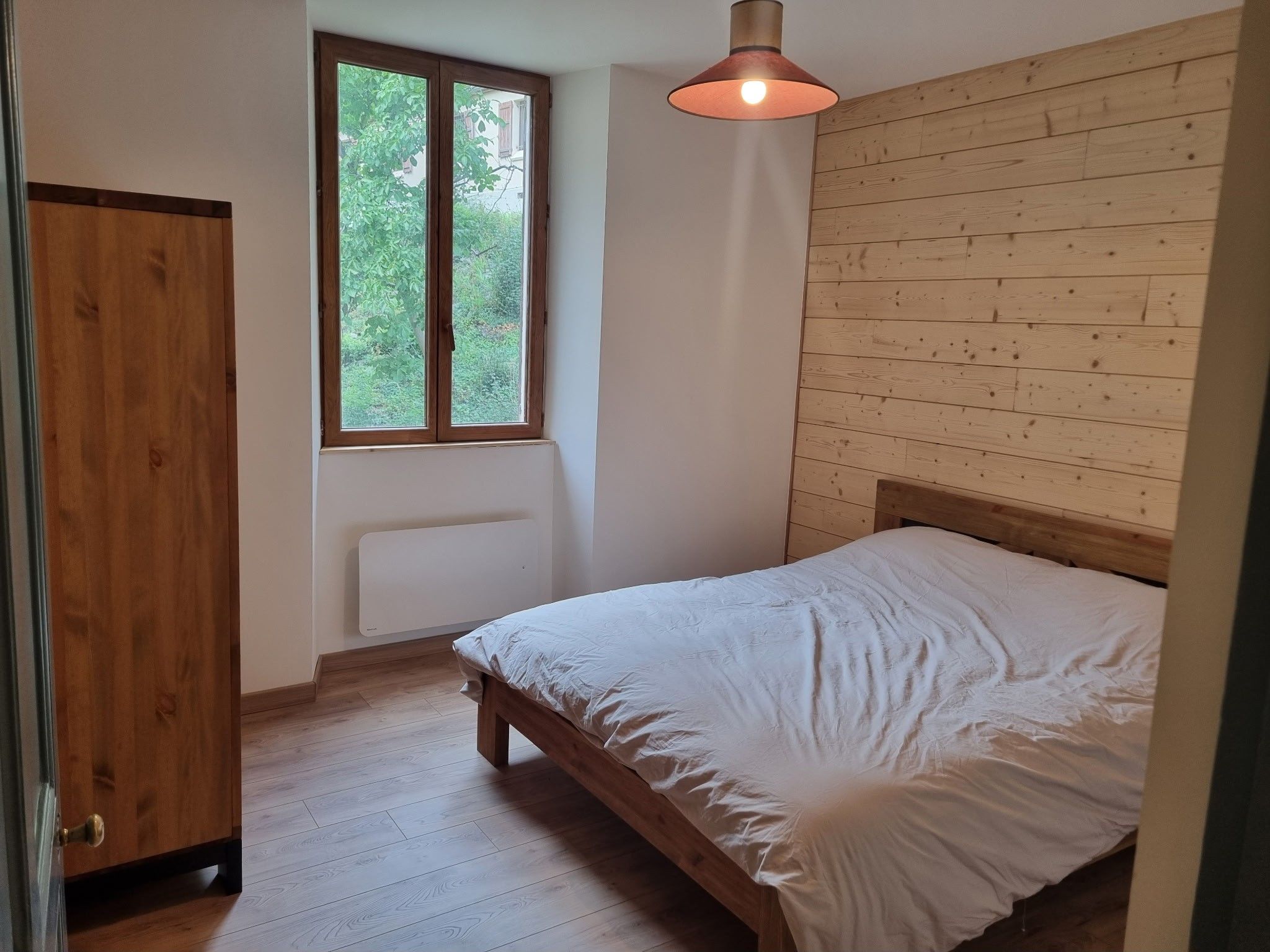 Une chambre avec un lit en bois, une fenêtre donnant sur un paysage verdoyant et une armoire en bois.