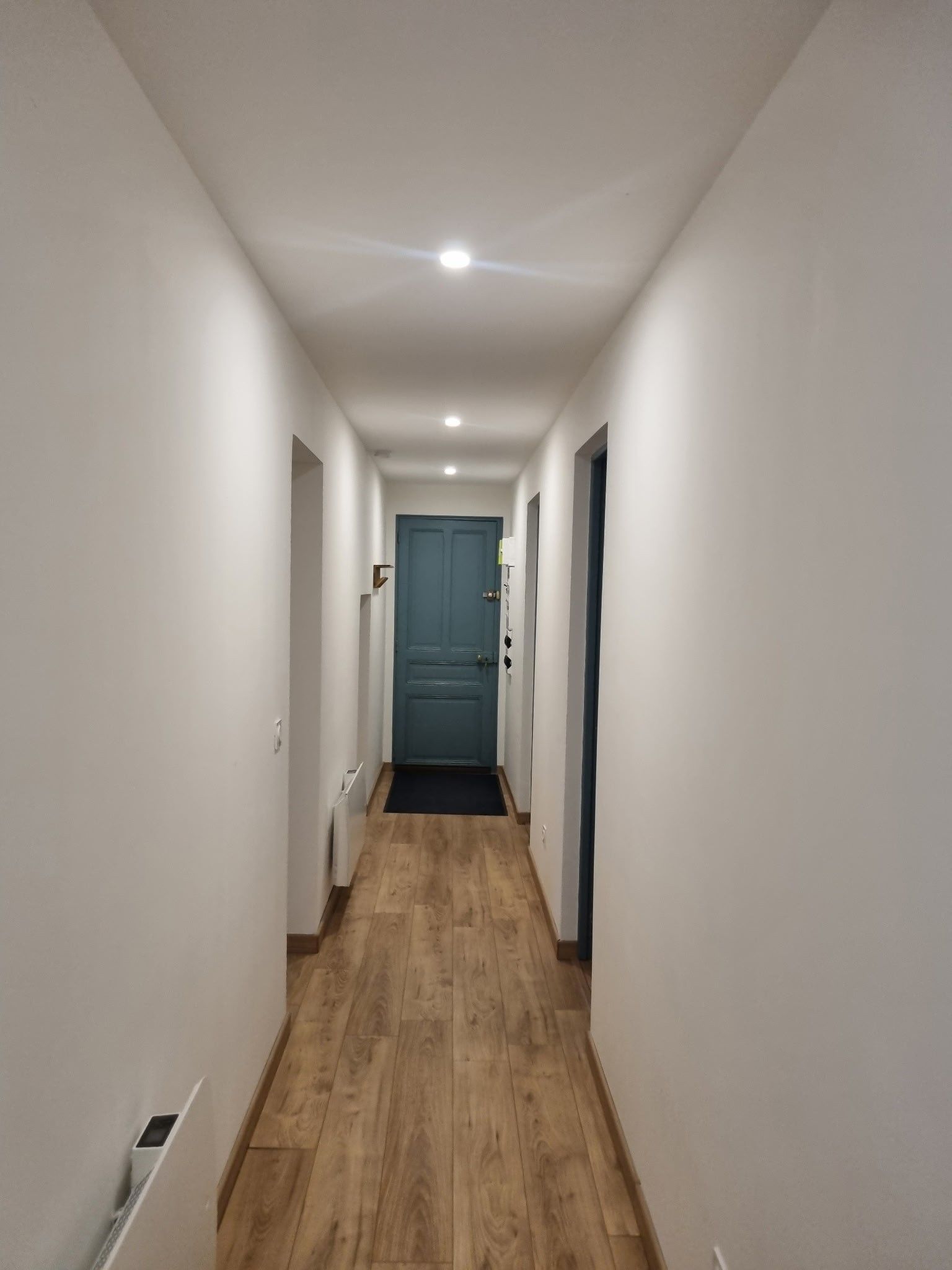 Un couloir étroit avec un plancher en bois, des murs blancs et une porte bleue au fond.