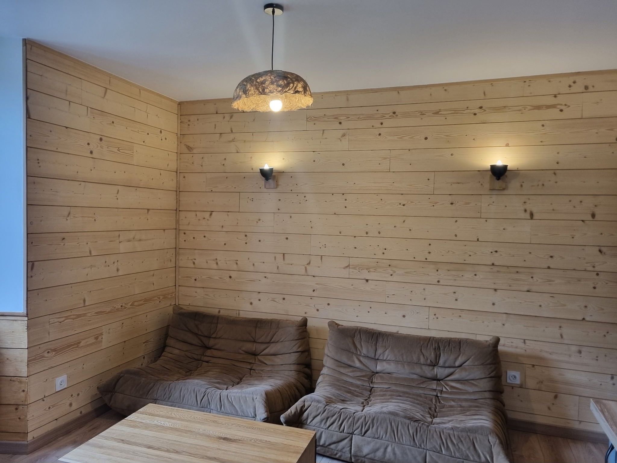 Habitación con paneles de madera, dos zonas de estar con cojines, una mesa de centro de madera y una lámpara colgante.