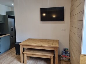 Een kleine keuken met een houten tafel en bankje, een tv aan de muur en een doos spelletjes op de vloer.