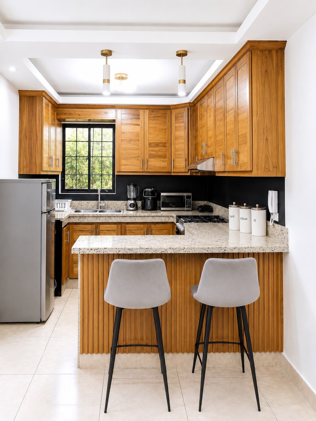 A modern kitchen with wooden cabinets, a granite countertop, and stainless steel appliances. There are two bar stools and a window providing natural light.