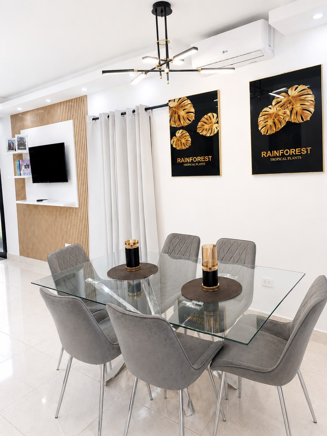 Un espace repas moderne avec une table en verre, des chaises à revêtement gris et un téléviseur à écran plat fixé au mur.