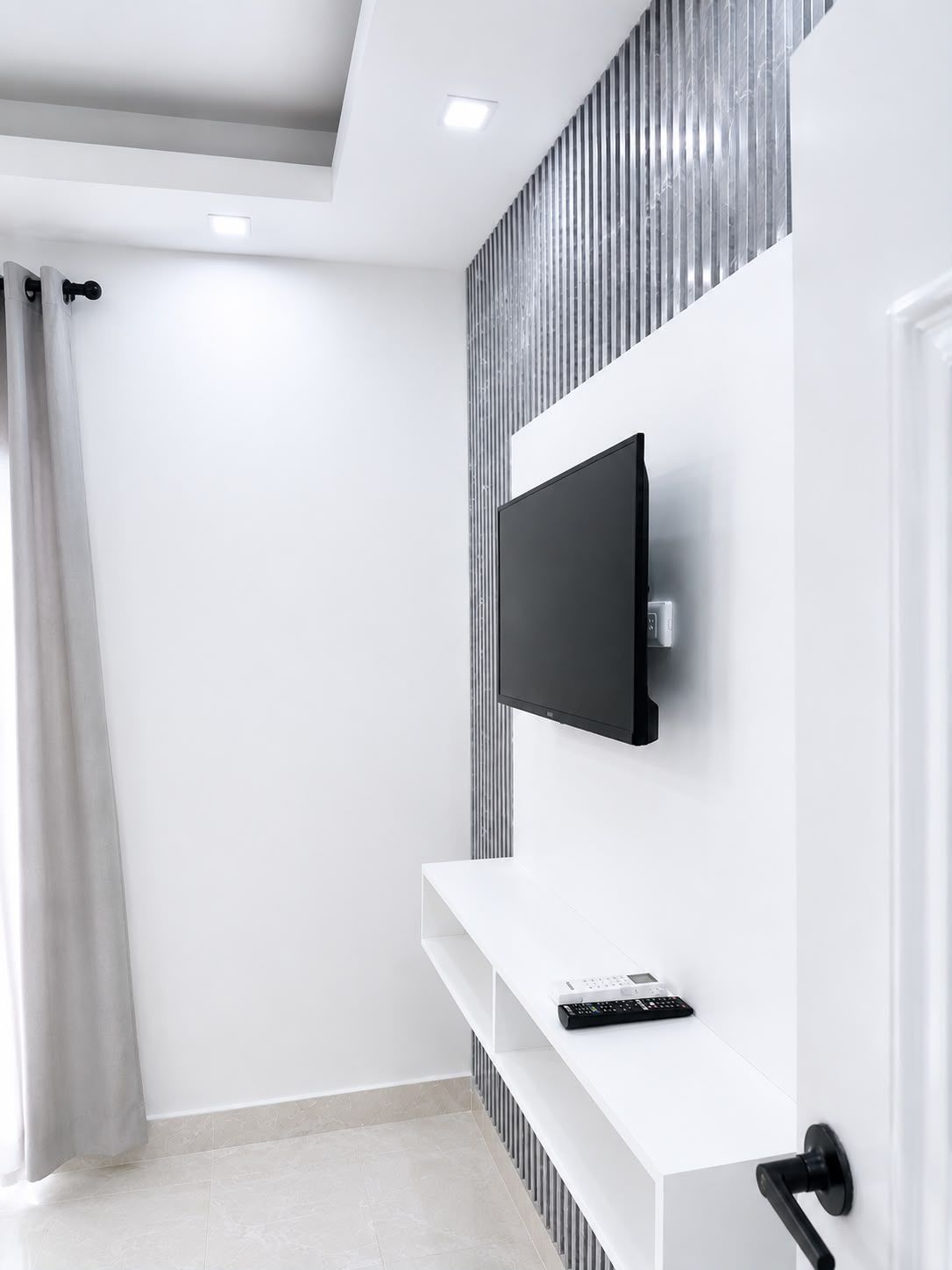 A modern living room with a flat-screen TV mounted on a wall, a white console table with a remote control, and a door with a black handle.