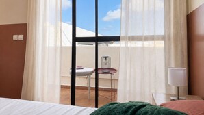 A hotel room with a bed, a nightstand, a lamp, a window with sheer curtains, and a view of a terrace with a chair and table. - The Olive Flat (Rabat)