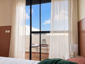 Habitación de hotel con cama, mesita de noche, lámpara, ventana con cortinas translúcidas y vista a una terraza con silla y mesa.