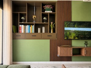 A living room with a TV mounted on a wall, a wooden entertainment unit with books and decorative items, and a window with curtains.