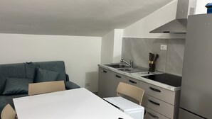 Eine kompakte Küchenzeile mit Spüle, Herd und Kühlschrank. Im angrenzenden Wohnbereich befindet sich ein Esstisch mit Stühlen sowie eine Couch.