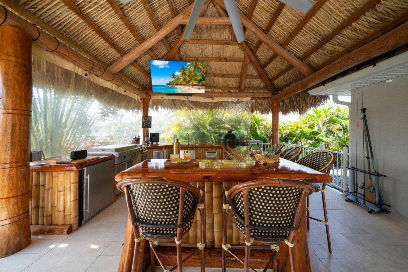 Une cuisine extérieure dotée d’un toit de chaume, avec un coin bar, des meubles en bambou et un écran de télévision diffusant des images d’un paysage tropical.