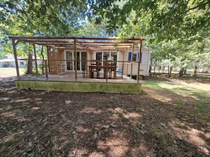 A covered outdoor area with a table and chairs, a wooden deck, and a mobile home in the background.