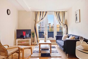 A modern living room with a flat-screen TV, a sofa, a coffee table, and a view of the city through large windows. - Stylish City Centre Living (Birmingham)