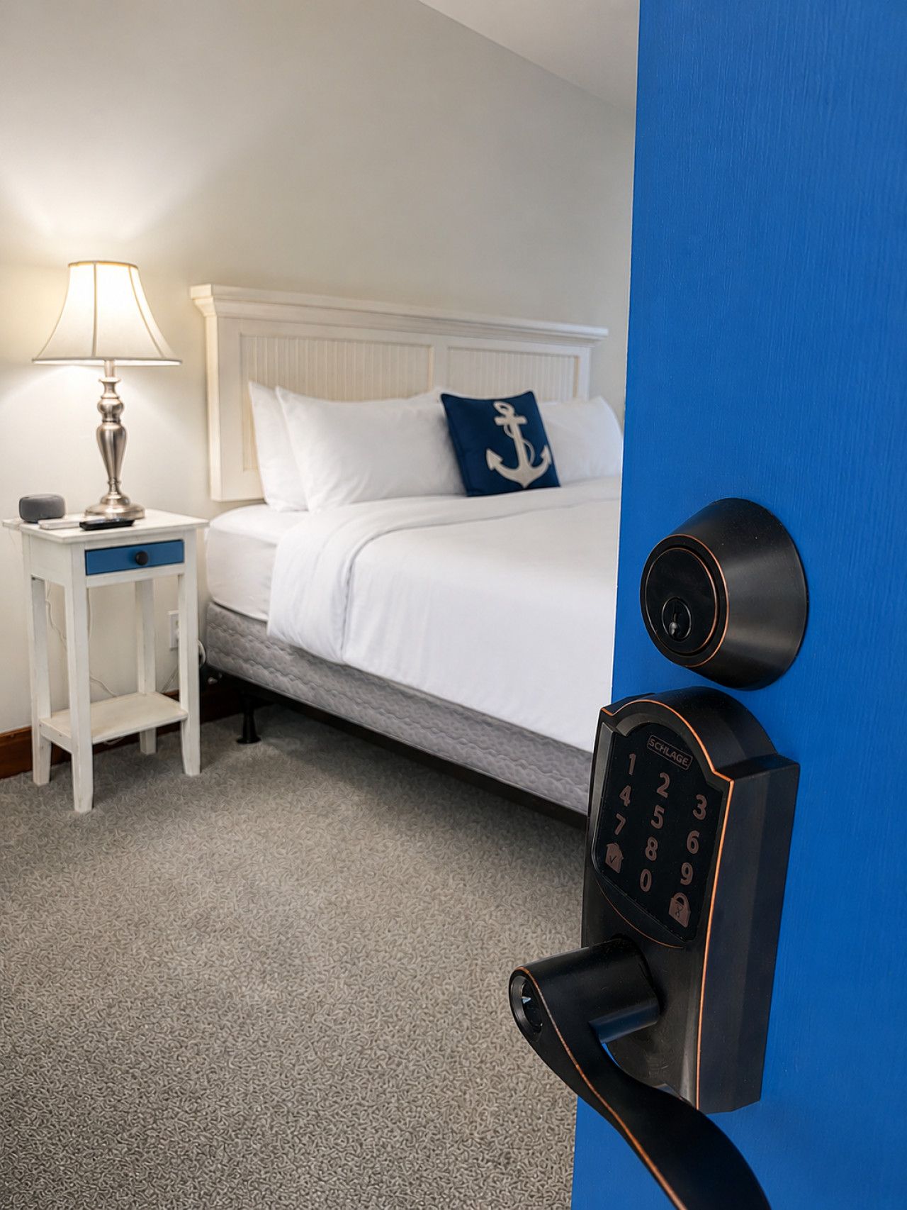 A hotel room with a bed, a nightstand, a lamp, and a modern electronic lock on the door.