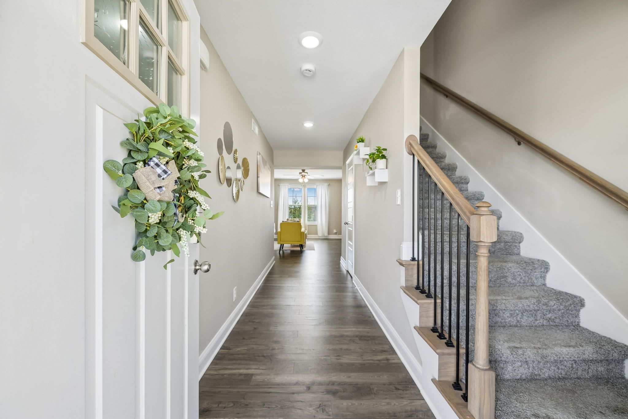 A hallway with a wooden staircase, a door with a wreath, and a dining area in the distance.
