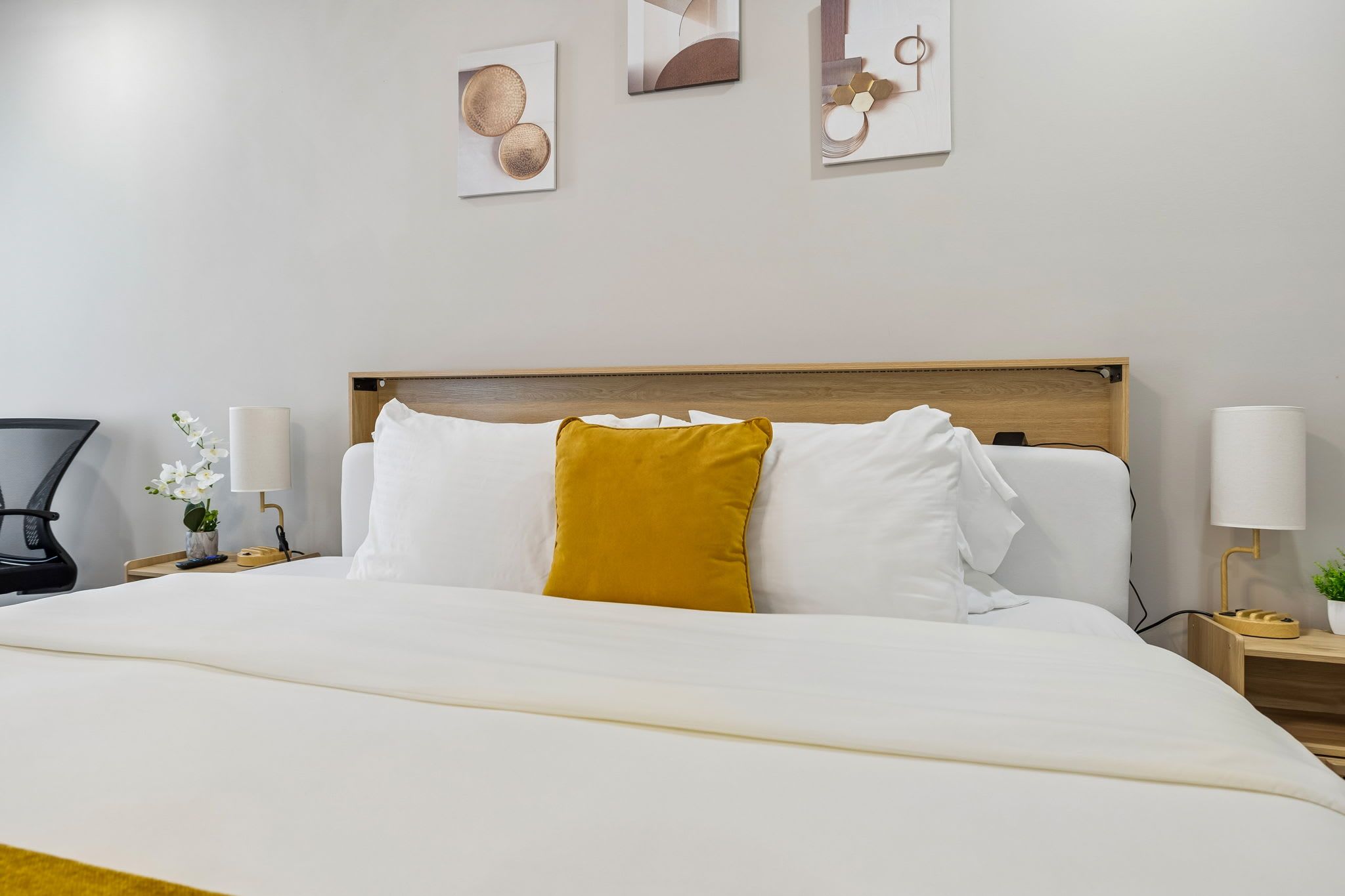 Un lit bien fait, avec du linge de lit blanc et un coussin de couleur moutarde, une table de chevet avec une lampe, et un mur orné d’œuvres d’art abstraites.