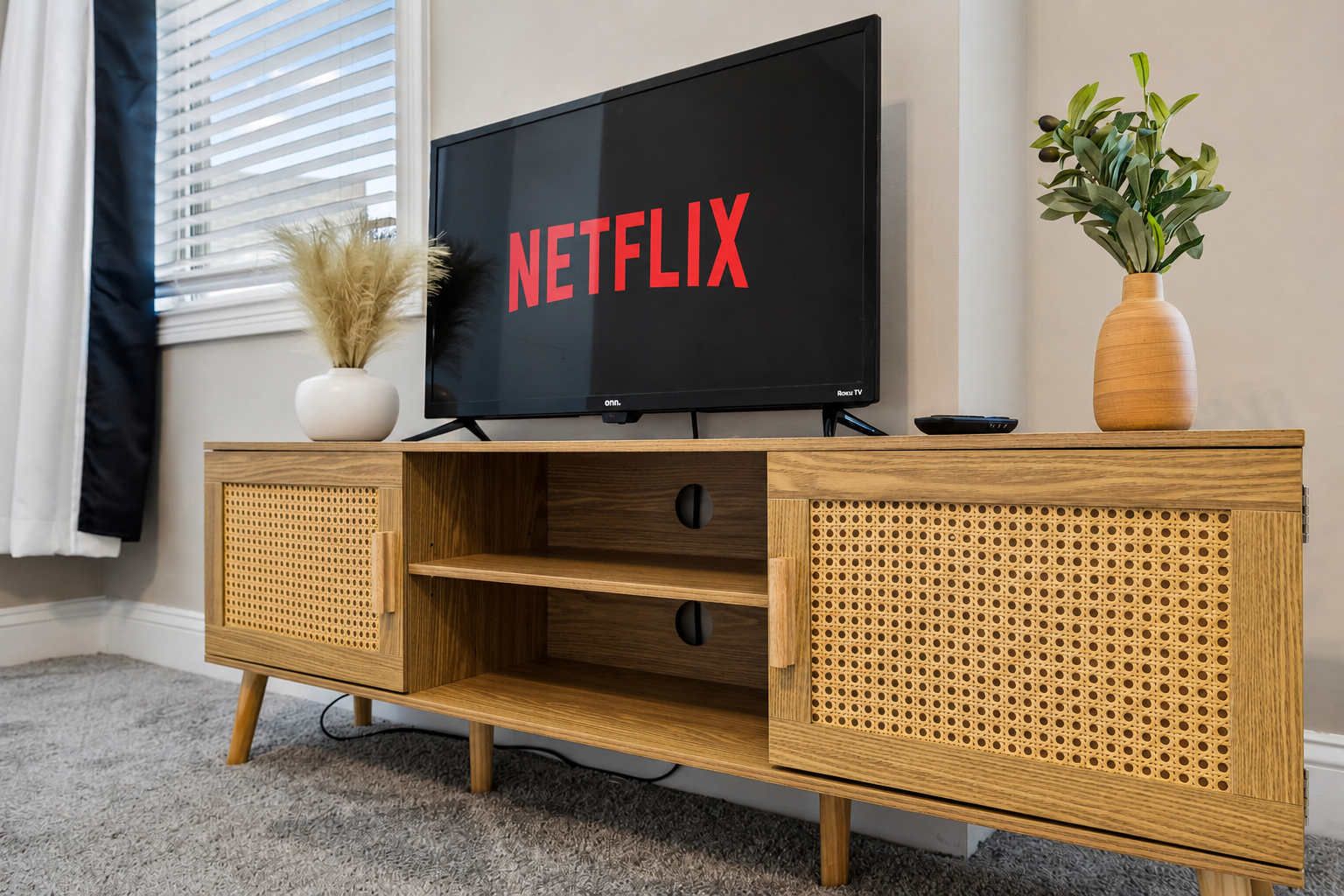 Un meuble de rangement en bois abritant un téléviseur à écran plat qui affiche le mot « NETFLIX » en lettres rouges, un vase blanc contenant des plantes séchées et un vase en bois avec des feuilles vertes.