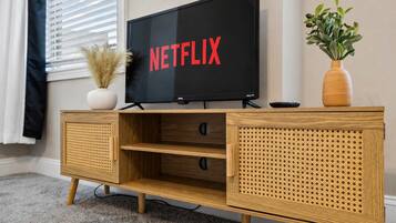 Un meuble de rangement en bois abritant un téléviseur à écran plat qui affiche le mot « NETFLIX » en lettres rouges, un vase blanc contenant des plantes séchées et un vase en bois avec des feuilles vertes.