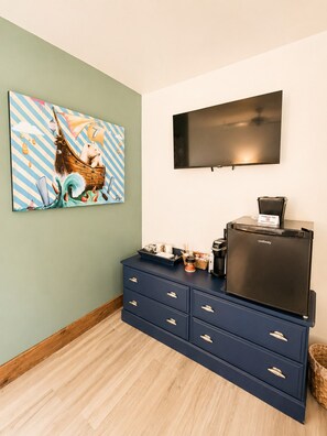 Una habitació amb televisió de pantalla plana penjada a la paret, un taulell blau amb calaixos, un quadre de disseny fantasiós i una cafetera sobre el taulell.