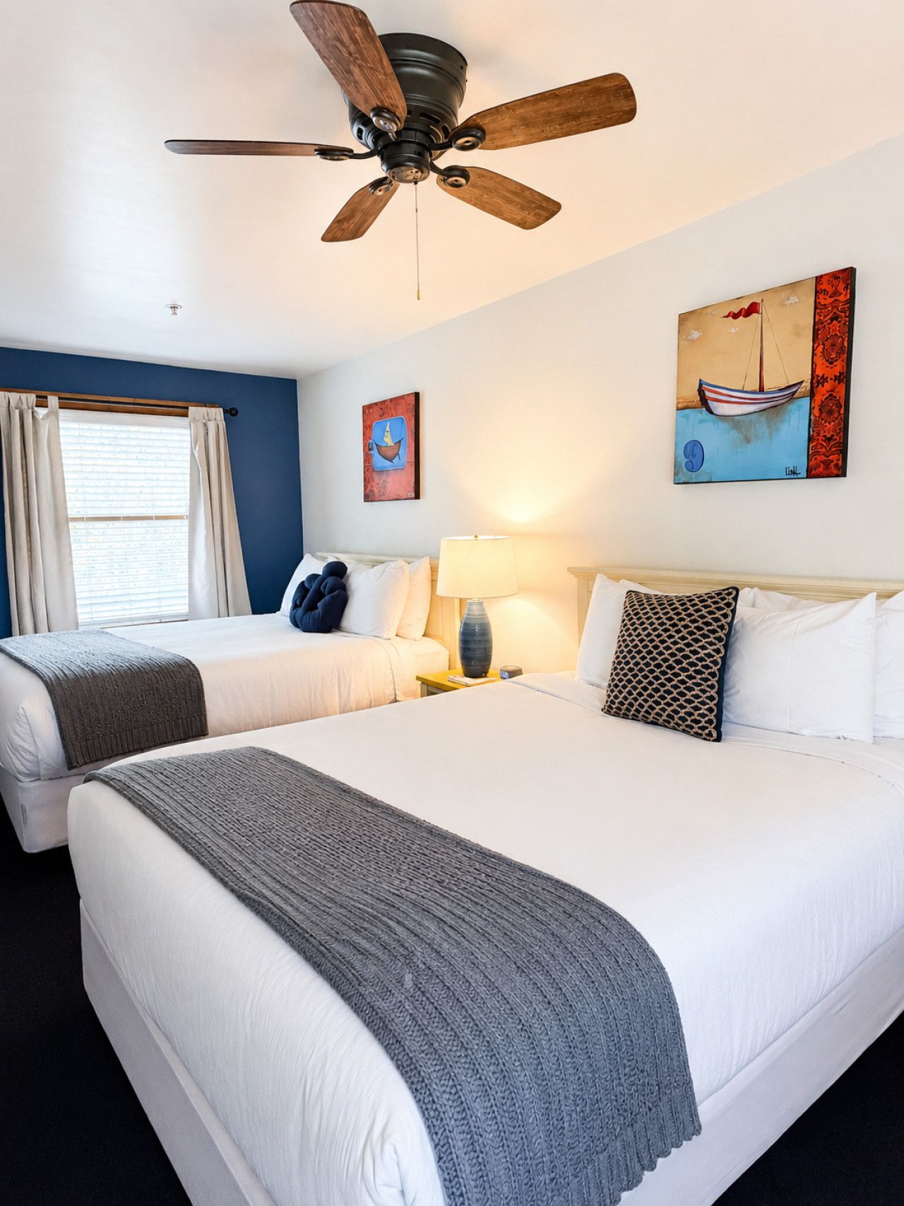 A hotel room with two beds, a ceiling fan, a window with curtains, and artwork on the walls.