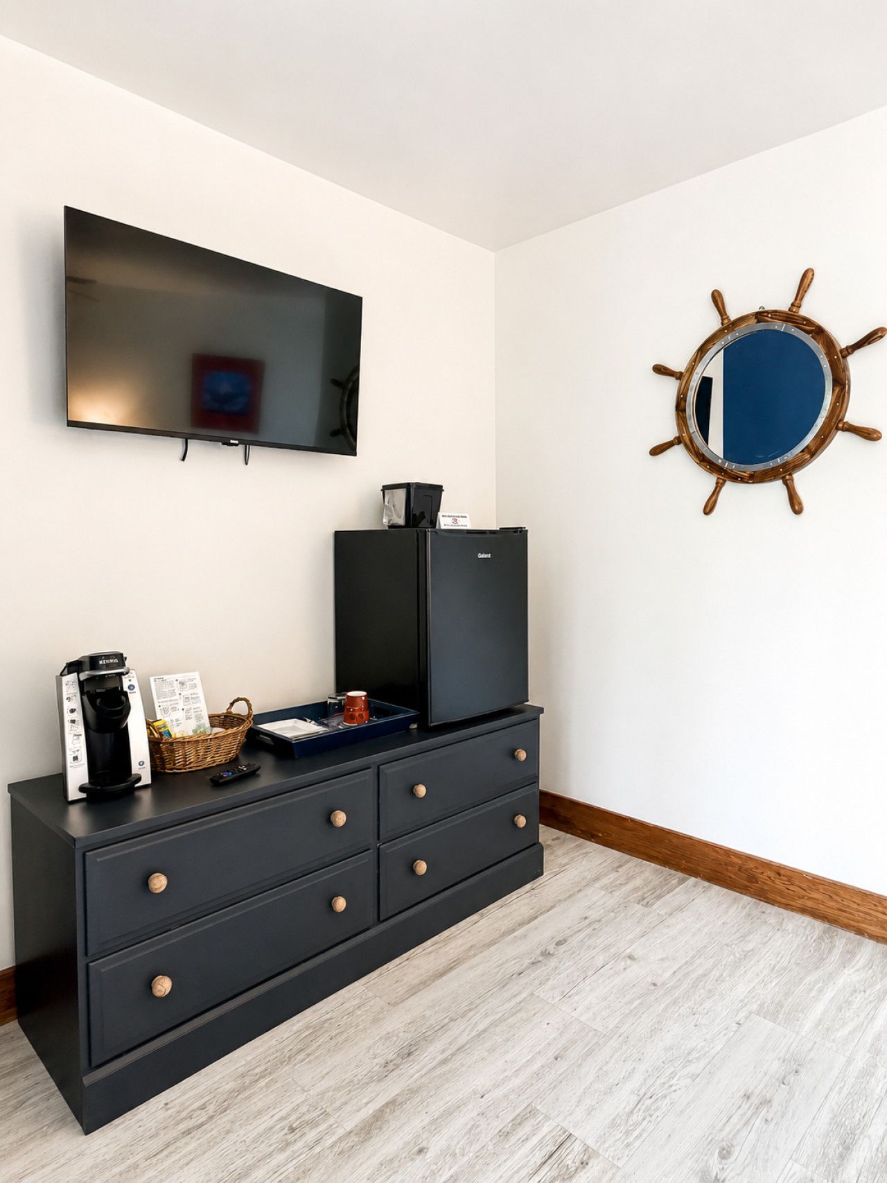 A room with a flat-screen TV mounted on the wall, a black entertainment unit with drawers, a coffee maker, a basket, and a ship's wheel decoration.