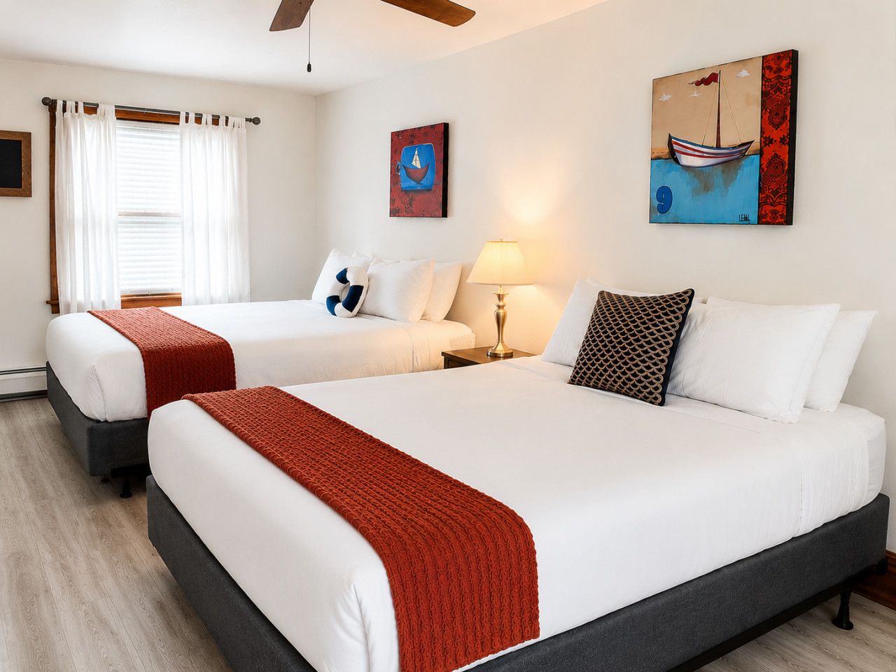 A hotel room with two beds, a window with curtains, a lamp, and artwork on the walls.